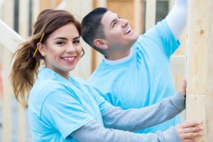 Hispanic couple work on house together that is being built for charity. The woman is holding a piece of wood while the man reaches to either hammer or measure the wood.